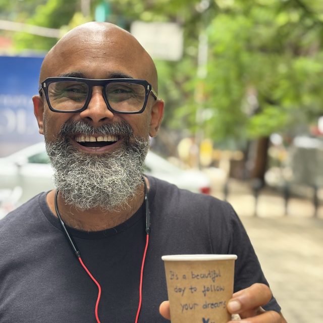 Venky outdoors, holding a paper cup of coffee, smiling at the camera.